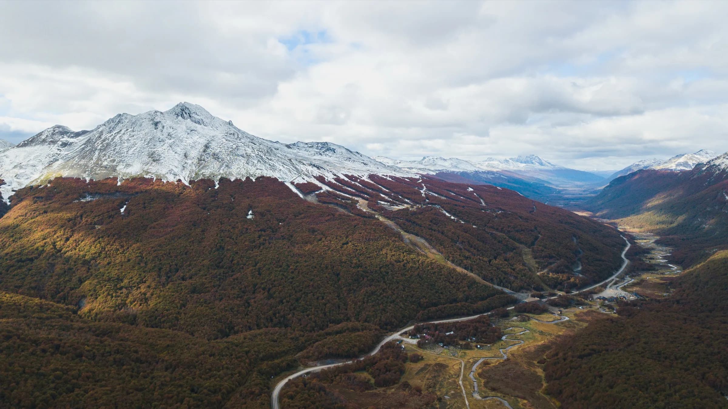 Vista aérea del valle en otoño, con el bosque rojizo y las cumbres con nieve